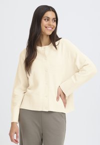 Grietos spalvos megztinis su rausvu raštu, apvalia iškirpte ir mygtukų uždarymu. Dėvimas virš šviesiai pilkų kelnių, su ilgomis rankovėmis ir laisvu siluetu.