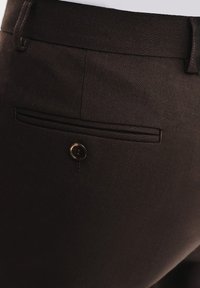 Brązowe spodnie z tkaniny o gładkiej fakturze, z kieszenią z lamówką i akcentem w postaci guzika z tyłu. Prosty design i dopasowany krój.