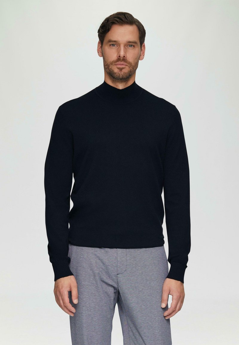 Schwarzer Rollkragenpullover aus weichem Strickmaterial, mit langen Ärmeln und figurnahem Design, kombiniert mit grauen, strukturierten Hosen.