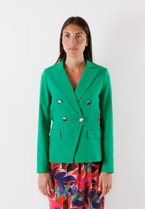 Blazer verde doppiopetto realizzato in tessuto liscio con bottoni argentati, revers a punta e due tasche frontali, indossato sopra un vestito a fantasia colorata.