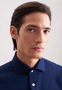 Camicia blu navy con colletto strutturato, chiusura con bottoni e tessuto testurizzato. Design liscio e aderente con bottoni bianchi visibili.