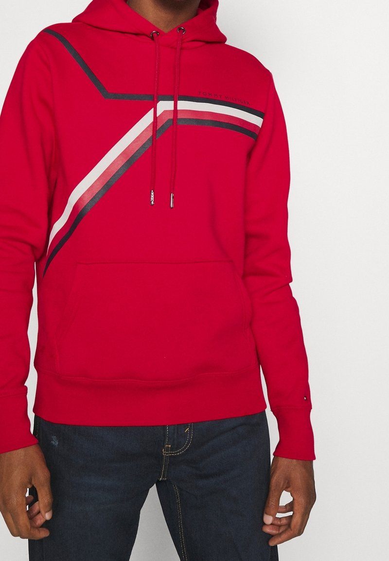 Sweat à capuche rouge avec des bandes diagonales noires, blanches et rouges sur la poitrine, poche avant et cordons, porté avec un jean foncé.