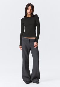 Kobieta stoi ubrana w czarną, dopasowaną bluzkę z długim rękawem oraz luźne szare spodnie, w czarnych butach, na jednolitym białym tle.