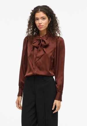 Brown satin blouse with a large bow at the collar, long sleeves with buttoned cuffs, paired with high-waisted black trousers.