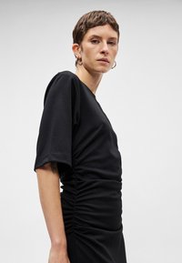 Μαύρο φόρεμα με εφαρμοστή σιλουέτα, κοντά μανίκια, λεπτομέρεια με ραφή στο πλάι και μαλακό βαμβακερό υλικό. Απλός σχεδιασμός χωρίς ορατά μοτίβα.