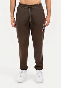 Braune Jogginghose aus weichem Stoff mit elastischem Bund und Kordelzug, Seitentaschen und einem vorderen Logo-Patch.