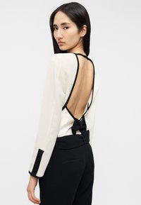 Μπλούζα με μακριά μανίκια σε κρεμ χρώμα με βαθύ άνοιγμα στην πλάτη και μαύρη επένδυση, διακοσμημένη με φιόγκο στη μέση. Συνδυάζεται με μαύρο, κομψό παντελόνι.