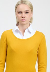 Gelbes Strickpullover mit langen Ärmeln und weitem Ausschnitt, über einem weißen Hemd mit Kragen getragen. Weiche Textur, lässiges Design.