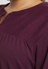 Mørkerød top med et tekstureret, mønstret skæring og en rund halsudskæring. Stoffet er blødt med en glat overflade og er samlet ved taljen.