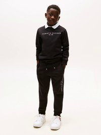 Crna dukserica s bijelim okvirom, logo "TOMMY HILFIGER", uparena s crnim trenirka hlačama i bijelim tenisicama, prikazuje minimalistički dizajn.