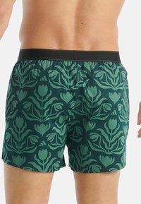 Muž v tmavě zelených šortkách se světle zeleným květinovým vzorem a černým páskem, zobrazený zezadu.