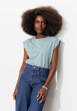 Femme aux cheveux afro naturels portant un haut bleu clair sans manches, un jean rayé bleu taille haute, un collier et un bracelet argentés, debout.