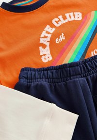 Orange T-Shirt mit dem Text „SKATE CLUB“ und Regenbogenstreifen; marineblaue Hose mit elastischem Bund; beiger Ärmelbesatz auf der linken Seite.