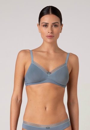 Donna con capelli scuri indossa un reggiseno azzurro chiaro e biancheria intima coordinata con bordo trasparente, in piedi davanti a uno sfondo pallido e uniforme.