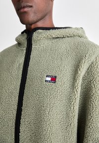 Fuzzy olijfgroene hoodie met ritssluiting, voorzien van een zwarte bies en een klein rechthoekig "TOMMY JEANS" logo in rood, wit en blauw.