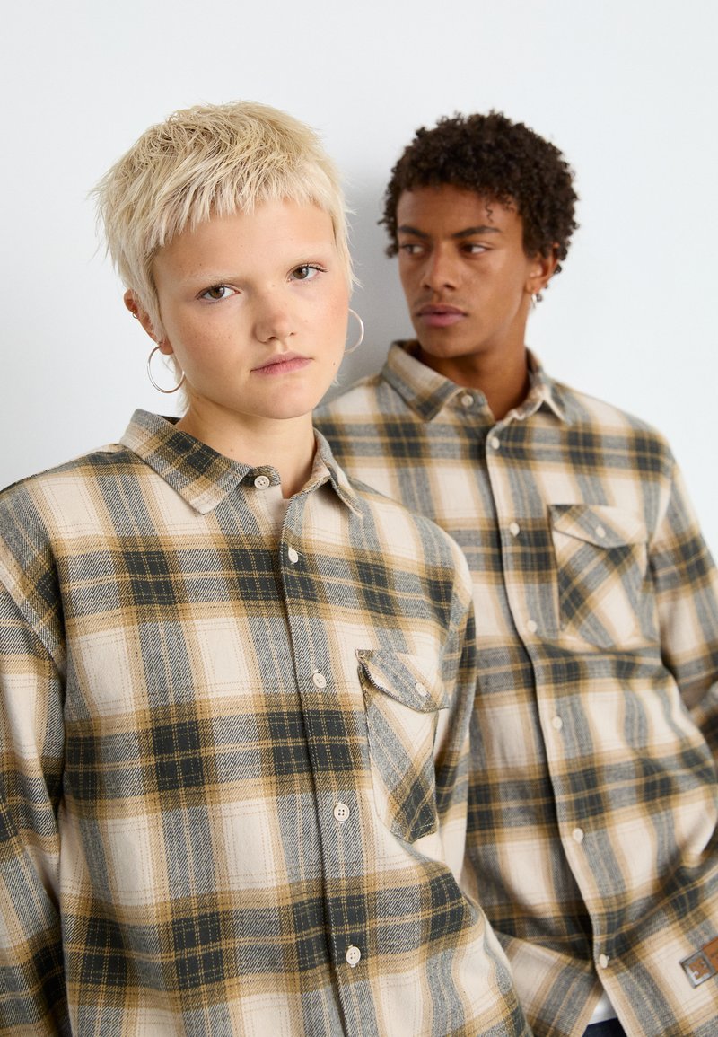 Koszule flanelowe w kratę w kolorach beżowym, zielonym i brązowym; długie rękawy, zapięcie na guziki, kieszeń na piersi i fakturowany materiał.