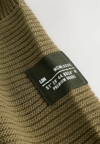 Khaki pletená látka s textúrovaným povrchom, s tmavozelenou nálepkou zobrazenou s koordinátmi a textom, našitou na boku.