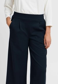 Pantaloni larghi navy con una texture morbida, dotati di vita alta e pieghe frontali. Le tasche sono visibili su entrambi i lati.