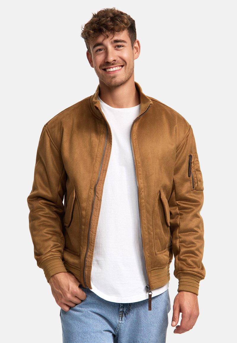 Veste aviateur marron en tissu doux avec fermeture éclair, deux poches latérales et poignets côtelés. Portée sur un t-shirt blanc et un jean bleu.