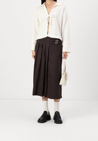 Bílé zkrácené svetr se zavázaným předním dílem, hnědá plisovaná sukně s přezkovými detaily, černé kožené boty a malá béžová kabelka.