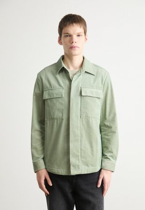 Green corduroy shirt with a collar, two chest pockets, and long sleeves, textured fabric, and straight hem, paired with dark pants.