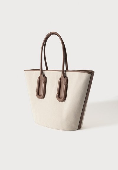 Μπεζ τσάντα ώμου με καφέ δερμάτινες λαβές και περιμετρικά, με επενδυμένες δερμάτινες βάσεις λαβών σε απλό φωτεινό φόντο.