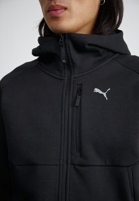 Svart hettejakke med glidelås laget av mykt stoff, med høy krage, frontlomme med glidelås, og en liten reflekterende Puma-logo på brystet.
