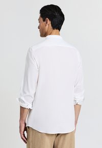 Mann, der ein weißes Langarmshirt mit aufgerollten Bündchen und beigen strukturierten Hosen trägt, von hinten vor einem schlichten Hintergrund gezeigt.