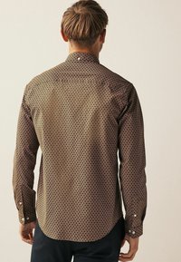 Camisa marrón de botones con un patrón circular repetido, de manga larga y botones blancos. Textura suave con un dobladillo curvado.