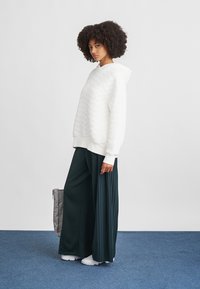 Felpa bianca con texture e vestibilità morbida, abbinata a pantaloni a gamba larga e plissettati verde scuro e sneakers bianche, completata da una borsa grigia.