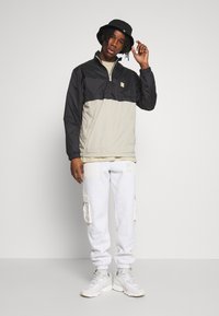 Giacca a vento leggera nera e beige con mezza cerniera, abbinata a pantaloni jogger grigi con tasche cargo e sneaker bianche.
