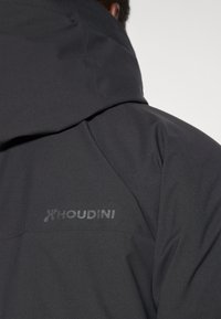 Czarna kurtka softshell z kapturem, z subtelnym logo Houdini na ramieniu. Gładka tekstura i elegancki, minimalistyczny design.