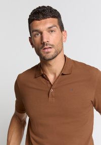 Bruine polo gemaakt van katoen, met een kraag, drieknoopsluiting en een klein logo op de borst. Korte mouwen en een slim-fit ontwerp.