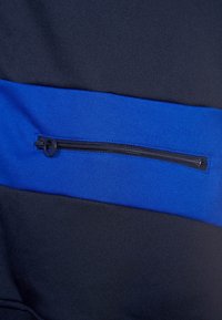 Svart tyg med en blå horisontell panel som har en dragkedjeficka. Mjuk textur med detaljer i funktionell metallkedja.