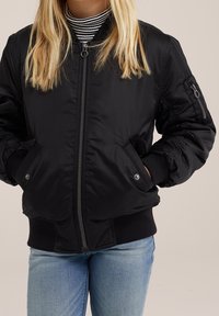 Blonde person wearing a black zip-up bomber jacket with pockets and a striped turtleneck, standing with hands in pockets.