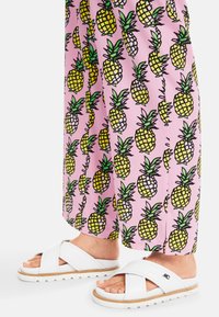 Weiße Rutschsandalen mit einem Kreuzriemen-Design auf einer strukturierten Sohle, kombiniert mit rosa Hosen, die ein sich wiederholendes Ananas-Muster in Gelb und Grün aufweisen.