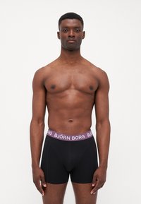 Schwarze Boxershorts mit lila Bund und "BJÖRN BORG"-Logo. Aus weichem Stoff gefertigt, präsentiert an einem Modell vor einem weißen Hintergrund.