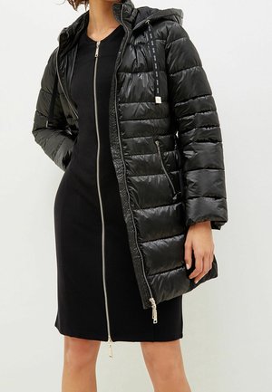 Kvinde iført en knælang sort kjole med lynlås og en sort dunjakke med hætte, hænderne i lommerne.