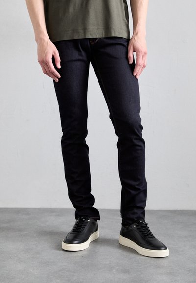 Jean en denim foncé avec une coupe slim et des coutures visibles, associés à des sneakers en cuir noir avec une semelle blanche.