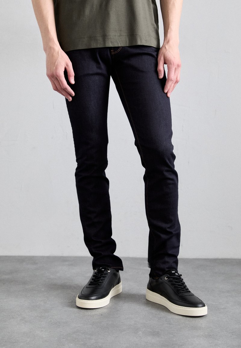 Jean en denim foncé avec une coupe slim et des coutures visibles, associés à des sneakers en cuir noir avec une semelle blanche.