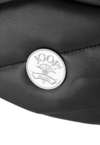 Borsa a mano in pelle nera con un prominente emblema circolare argento recante il testo "JOOP! JEANS" e un motivo decorativo. Texture liscia e cuciture dettagliate.