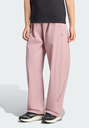 Persona che indossa pantaloni rosa larghi, top nero e sneaker bianche e nere in piedi davanti a uno sfondo semplice.