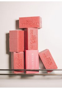 Six barres de savon rectangulaires roses empilées sur une étagère en verre ; cinq barres sont embossées du texte "Sitre Horosoaps", une est emballée et étiquetée "Better Wettie".