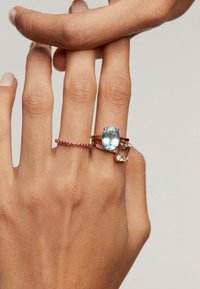 Tres anillos en una mano: una delgada banda de gema roja, una gran piedra ovalada azul y una piedra rectangular clara, todos engastados en oro rosa.