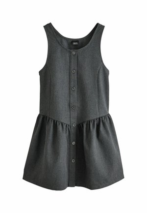 Ein junges Mädchen steht und trägt ein weißes langärmeliges Hemd unter einem dunkelgrauen, geknöpften Kleid, weiße Socken mit rotem Rand und schwarze Schuhe.
