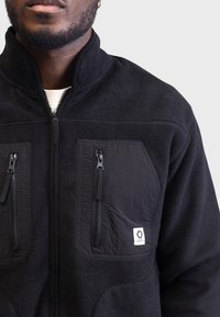 Černá fleecová bunda s vysokým límcem, obsahující dvě kapsy na zip na hrudi a nášivku s logem. Textura je měkká a teplá.