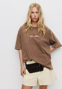 Barna oversize póló "SPICY PASSION" felirattal, sárga csíkos rövidnadrággal és fekete, studsos deréktáskával párosítva.