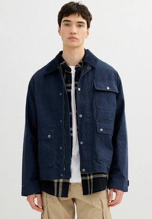 Joven con chaqueta abotonada azul marino sobre una camisa de cuadros negra y camiseta blanca, combinada con pantalones cargo beige, posando contra un fondo blanco.