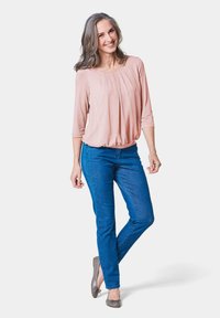 Helles rosa Blusenshirt mit ¾-Ärmeln und Falten, kombiniert mit blauen Slim-Fit-Jeans. Graue Ballerinas runden das Outfit ab. Schlichtes, sauberes Design.