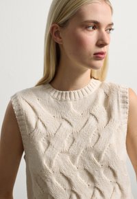 Junge Frau mit glattem, blondem Haar, die einen ärmellosen, cremefarbenen Pullover mit Zopfmuster trägt und nach rechts schaut, vor einem schlichten Hintergrund.